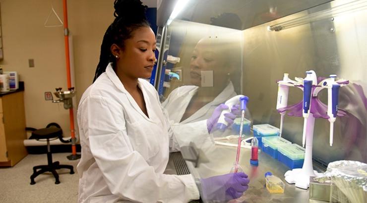 Nai Brown in a lab coat and sterile gloves standing on one side of a clear barrier while her hands perform a procedure on the other side of the barrier in a Delaware Tech laboratory