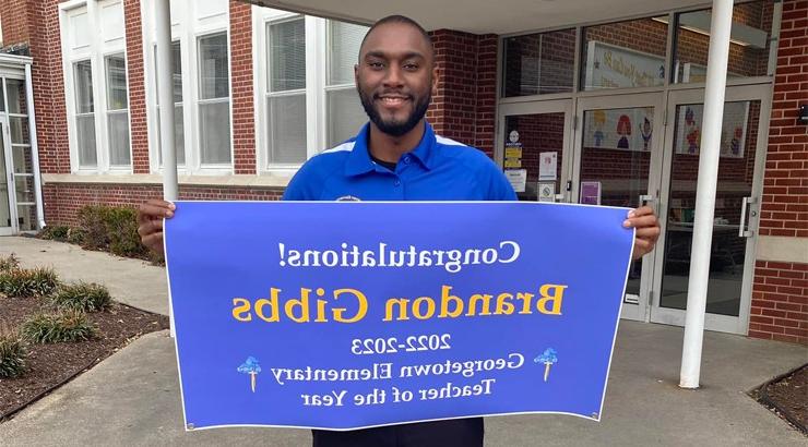 Delaware Tech Alumni Brandon Gibbs standing in front of Georgetown Elementary School holding a sign that says 